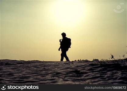 Silhouette of modern infantry soldier in tactical ammunition, armed with service rifle, standing in sandy, desert area on sunset or sunrise background. Army ranger patrolling territory at dawn. Silhouette of modern army soldier in dawn patrol