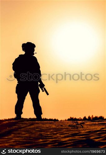 Silhouette of modern infantry soldier in tactical ammunition, armed with service rifle, standing in sandy, desert area on sunset or sunrise background. Army ranger patrolling territory at dawn. Silhouette of modern army soldier in dawn patrol