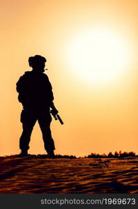 Silhouette of modern infantry soldier in tactical ammunition, armed with service rifle, standing in sandy, desert area on sunset or sunrise background. Army ranger patrolling territory at dawn. Silhouette of modern army soldier in dawn patrol