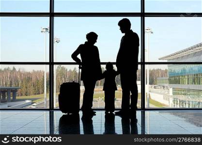 silhouette of mother, father and daughter with luggage standing near window in airport