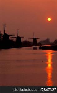 Silhouette of old-fashioned, wooden windmills along canal with yellow setting sun in background