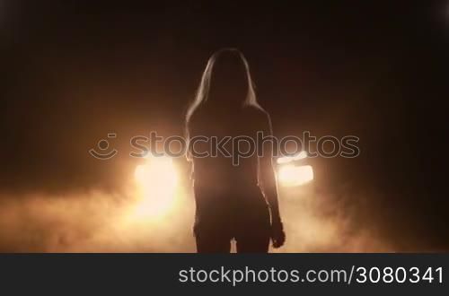 Silhouette of sensual joyful woman dancing in night time over bright car headlights background. Beautiful girl in shorts standing in clouds of smoke, making dance moves in yellow headlights of car at night. Slow motion.