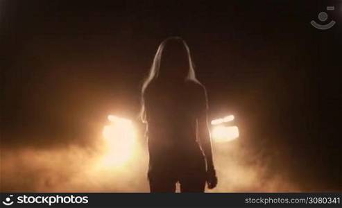 Silhouette of sensual joyful woman dancing in night time over bright car headlights background. Beautiful girl in shorts standing in clouds of smoke, making dance moves in yellow headlights of car at night. Slow motion.