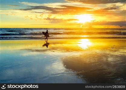 Silhouette of surfer walking on sandy beach with surfboard, sunset skyline over ocean waves, Bali, Indonesia