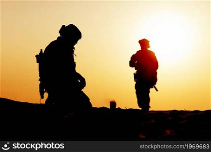 Silhouette of tired army soldiers or military contractors in tactical ammunition, returning from patrol, sitting on knees on background of desert sunset, heating drink after hard mission or battle. Silhouettes of soldiers resting after night watch