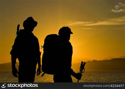 Silhouette of two adventurers admiring the radiant sunset over a lake with hills and forests in the background