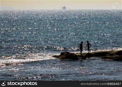 Silhouette of Two Children Playing on Reef near Sea at Sunset and Supply Ship in background
