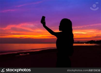 Silhouette of woman taking a photo of sunset with mobile phone, at sea beach