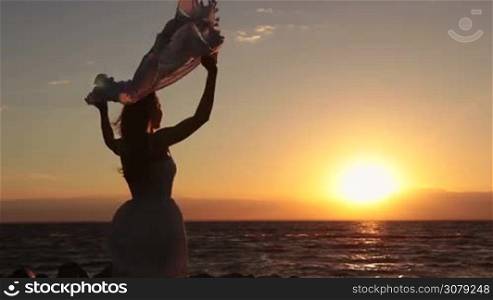 Silhouette of young blonde female with flying scarf fluttering in wind standing on seashore while enjoying freedom, feeling happy and tranquil at beach at sunset. Carefree girl holding silk scarf on seaside in rays of setting sun.