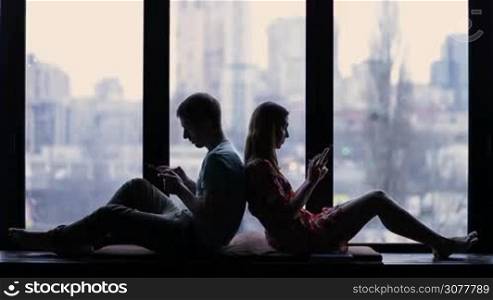 Silhouette of young couple sitting back to back at home and using smartphones against window background. Internet addicted couple using mobile phones, absorbed in social networks. Concept of apathy connected to the alienation due to new technologies.