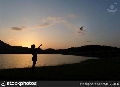 Silhouette of young woman playing a kite by the lake at sunset.