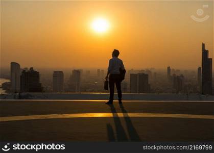 Silhouette Rear view of businessman standing with carrying the business bag and looking the vision over the cityscape background at sunset time with lens flare,Business success concept
