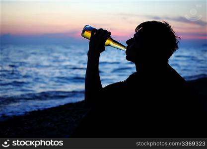 Silhouette teenager boy drinking beer on stone seacoast in evening