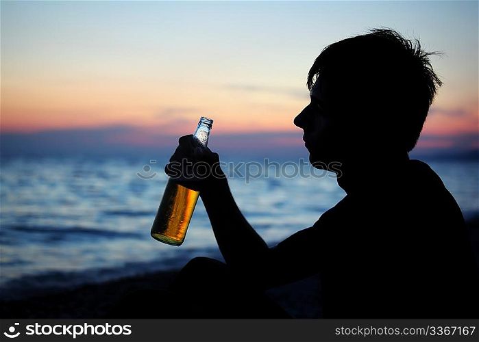 Silhouette teenager boy with beer bottler on stone seacoast in evening
