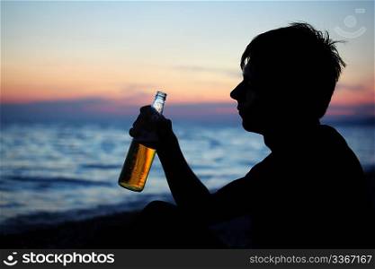 Silhouette teenager boy with beer bottler on stone seacoast in evening