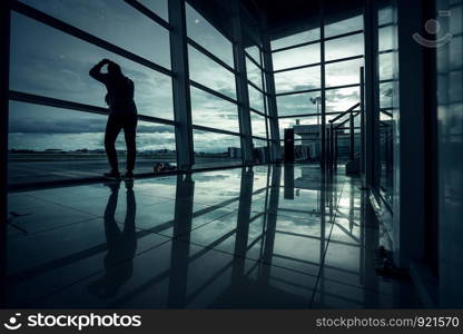 Silhouette young man is standing near window at the airport and watching plane before departure