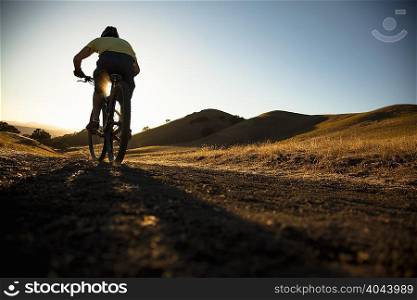 Silhouetted surface view of young man mountain biking up dirt track, Mount Diablo, Bay Area, California, USA