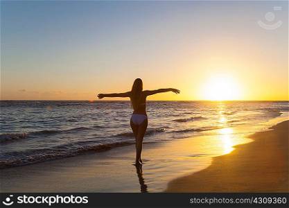 Silhouetted young woman with arms open on beach at sunset, Dominican Republic, The Caribbean