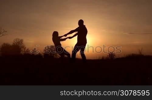 Silhouettes of couple in love holding by hands and spinning around in glowing of awesome sunset. Joyful couple sharing the best moments of togetherness, having fun while spending great time outdoors at sunset. Slow motion. Steadicam stabilized shot.