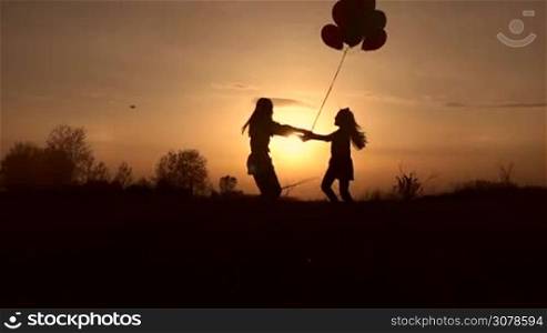Silhouettes of joyful mother and cute little daughter having fun in the field at sunset. Mother and little girl with colorful balloons holding hands and spinning around in the rays of setting sun while spending weekend in countryside. Slow motion.