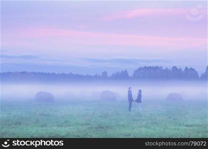 Silhouettes of man and woman standing opposite each other and holding hands in foggy field with haystacks at sunset