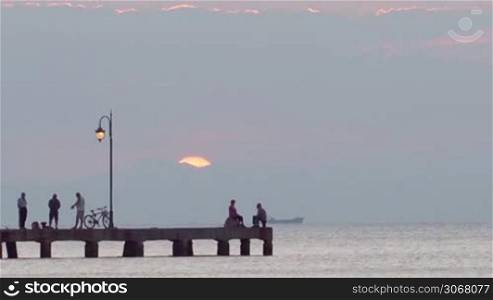 Silhouettes of people relaxing on a pier at sunset standing talking, fishing or riding a bicycle as the sun dips below the hills. Ship is passing by