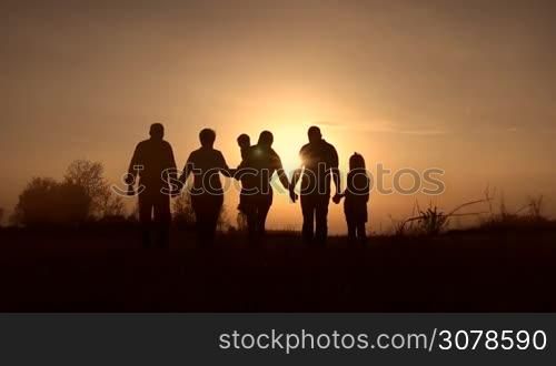 Silhouettes of three generation family holding hand in hands walking in the meadow in glowing of beautiful sunset. Rear view. Happy family with two kids enjoying amazing view of setting sun outdoors in spring. Slow motion. Steadicam stabilized shot.