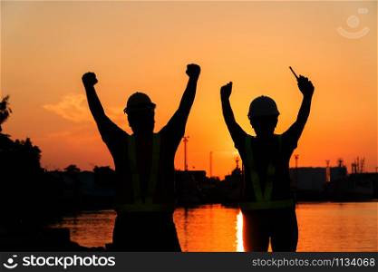 Silhouettes of worker and engineer Raising the hand up and standing on the shipyard. Background is oil storage silo. Teamwork cooperation And success in working together concept.