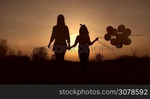 Silhouettes of young mother and her cute little daughter with colorful balloons holding hands walking in the meadow in the rays of setting sun. Positive family spending time together outdoors at sunset. Slow motion. Steadicam stabilized shot.