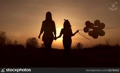 Silhouettes of young mother and her cute little daughter with colorful balloons holding hands walking in the meadow in the rays of setting sun. Positive family spending time together outdoors at sunset. Slow motion. Steadicam stabilized shot.