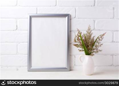 Silver frame mockup with grass and green leaves in pitcher vase . Silver frame mockup with meadow grass and green leaves in pitcher vase near painted brick wall. Empty frame mock up for presentation artwork. Template framing for modern art.