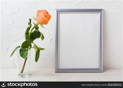 Silver frame mockup with orange-apricot rose in glass vase. Empty frame mock up for presentation artwork.. Silver frame mockup with orange-apricot rose in glass vase
