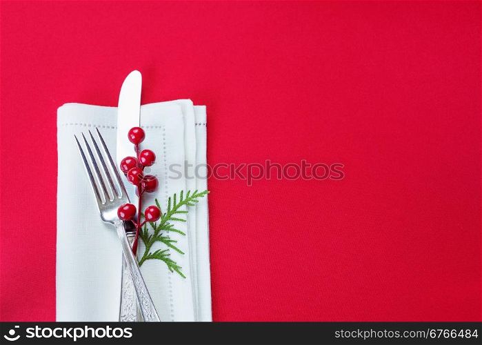 Silver knife and fork, and red holly berries and green thuja branch lie on the white linen napkin, which is located on a table covered with a red tablecloth, top view