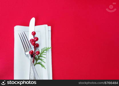 Silver knife and fork, and red holly berries and green thuja branch lie on the white linen napkin, which is located on a table covered with a red tablecloth, top view