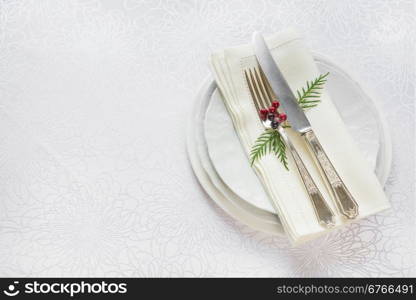 Silver knife and fork, and red holly berries and green thuja branches lie on the white porcelain plate, which is located on a table covered with a white tablecloth, top view