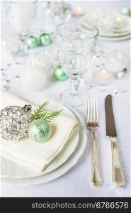 Silver knife and fork, beautiful wine glass, and green christmas balls lie on the white porcelain plate, which is located on a table covered with a white tablecloth