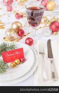 Silver knife and fork, christmas ball and green spruce branche lie on the white porcelain plate, glass of red wine, which is located on a table covered with a white tablecloth