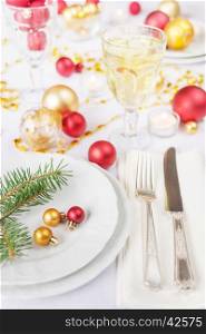 Silver knife and fork, christmas ball and green spruce branche lie on the white porcelain plate, glass of white wine, which is located on a table covered with a white tablecloth
