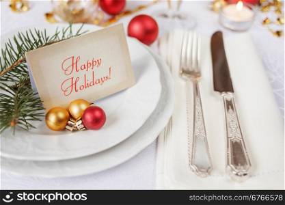 Silver knife and fork, christmas ball and green spruce branche lie on the white porcelain plate, which is located on a table covered with a white tablecloth