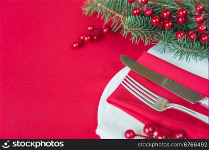 Silver knife and fork lie on the red linen napkin, as well as red holly berries and green spruce branch, which is located on a table covered with a red tablecloth, with space for text