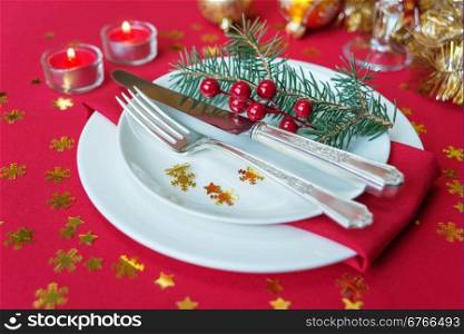 Silver knife, fork, dinner plates, holly berries, pine branch and burning candles, which is located on a table covered with a red tablecloth