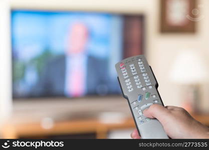 Silver modern TV remote control being pressed by thumb with out of focus screen background