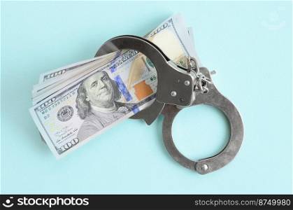 Silver police handcuffs and hundred dollar bills lies on light blue background.. Silver police handcuffs and hundred dollar bills lies on light blue background
