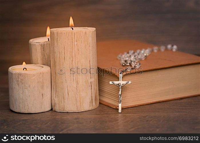 Silver rosary and crucifix resting on closed book near the candles on wooden table, religion school concept. Vintage style.