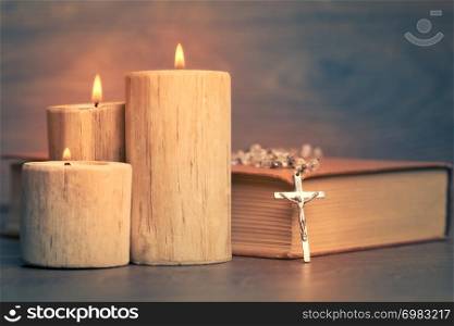 Silver rosary and crucifix resting on closed book near the candles on wooden table, religion school concept. Vintage style.