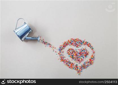 silver watering can pouring sugar baking colorful sprinkles in shape of heart on a light beige background.. silver watering can pouring sugar baking colorful sprinkles in shape of heart on light beige background.