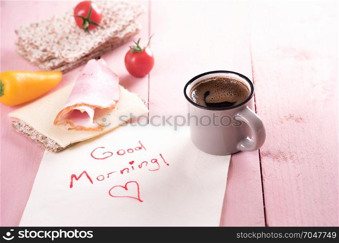 Simple, healthy meal with whole-wheat crispbread, tomatoes, cheese, meat and a cup of coffee, on a napkin with a good morning message, in the morning light.
