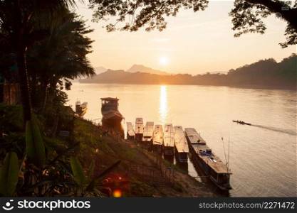 Simple life, golden light shines down around the Mekong River, small wooden boat returning home in the evening, vegetable garden and local boat foreground. Mountains and sunset background. Luang Prabang, Laos. The world heritage town.