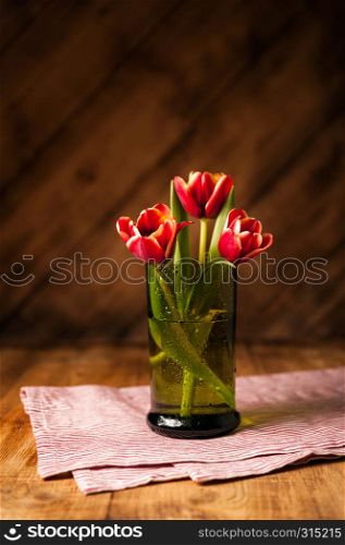 Simple rustic still life with blooming red tulips in a green glass on a wooden table with a striped napkin. Fine art floral background. Close Up.. Simple rustic still life with blooming red tulips in a green glass on a wooden table with a napkin.