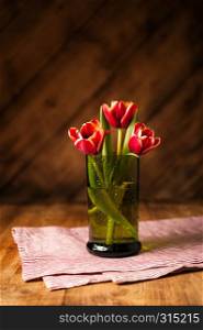 Simple rustic still life with blooming red tulips in a green glass on a wooden table with a striped napkin. Fine art floral background. Close Up.. Simple rustic still life with blooming red tulips in a green glass on a wooden table with a napkin.
